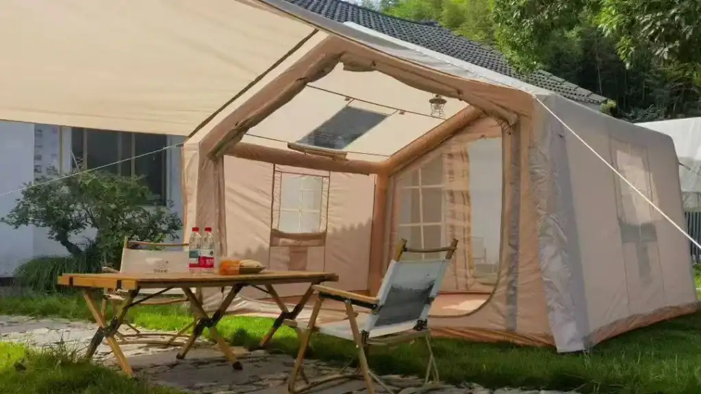 Une grande tente de camping beige avec des fenêtres transparentes et une table garnie de boissons et d'en-cas, entourée d'herbe verte et d'arbres.