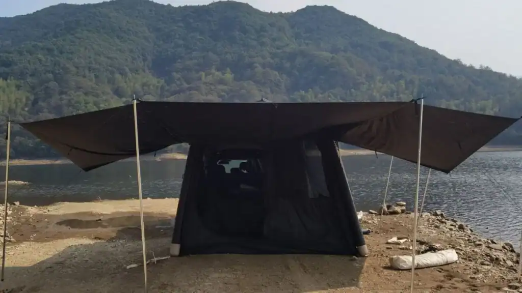 Camping tent set up by the waterfront with a large canopy extending over the front, overlooking a mountainous landscape.