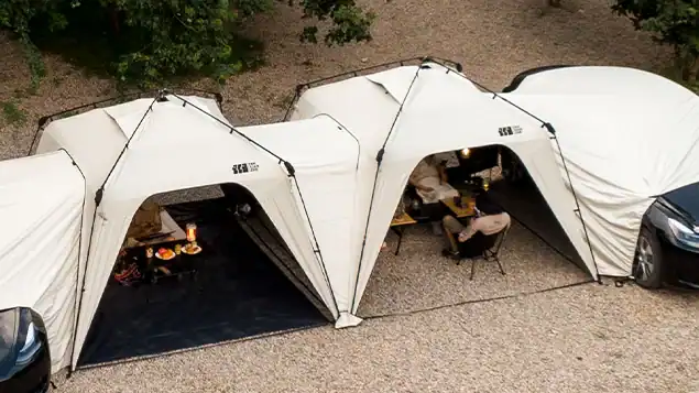 خيام تخييم بيضاء متعددة على جانب السيارة متصلة ببعضها البعض، لتشكل مناطق مظللة للمعيشة والنوم بجانب السيارات المتوقفة على الحصى