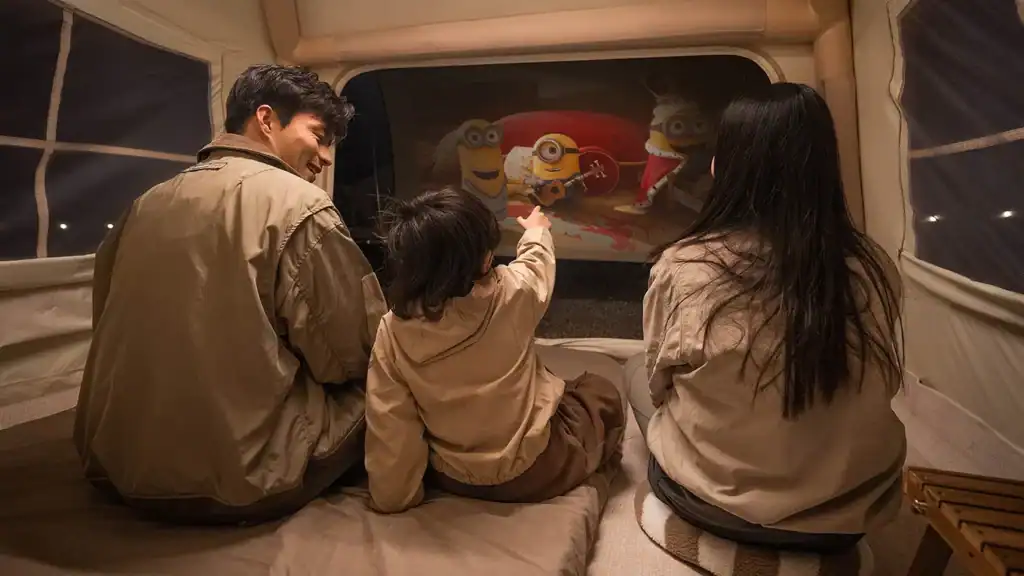 A family of three sits inside a spacious camping tent watching an animated movie on a portable screen, child pointing excitedly
