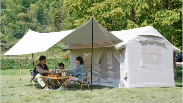 دليل إصلاح الخيام القابلة للنفخ: إصلاح التسريبات والصمامات 1 عائلة مكونة من ثلاثة أفراد يجلسون على طاولة تخييم خشبية تحت مظلة بيج اللون متصلة بخيمة كبيرة في الهواء الطلق محاطة بالأشجار الخضراء