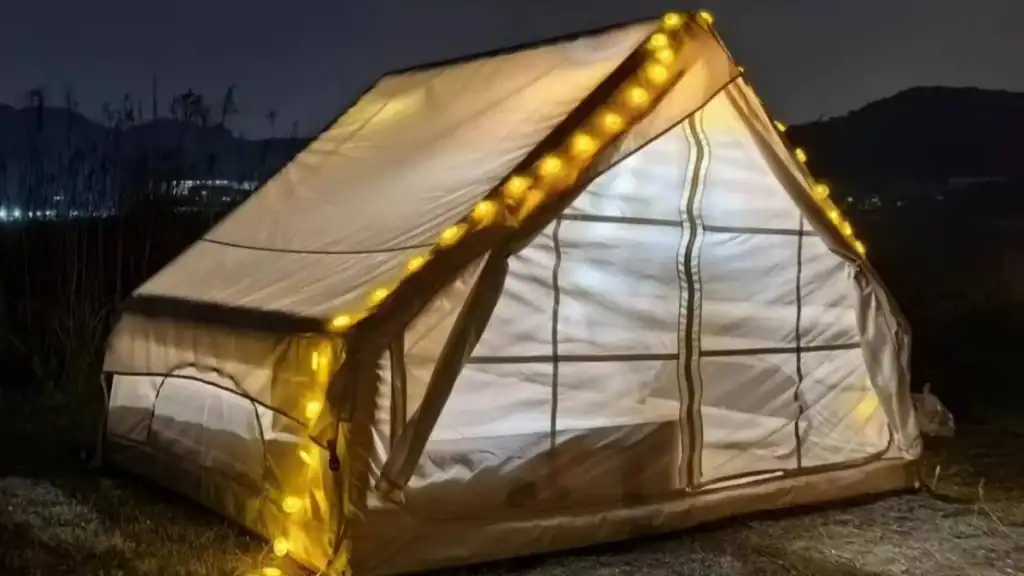 A beige camping tent illuminated from inside with soft lights, decorated with a string of warm yellow bulbs along its edges in an outdoor setting at night
