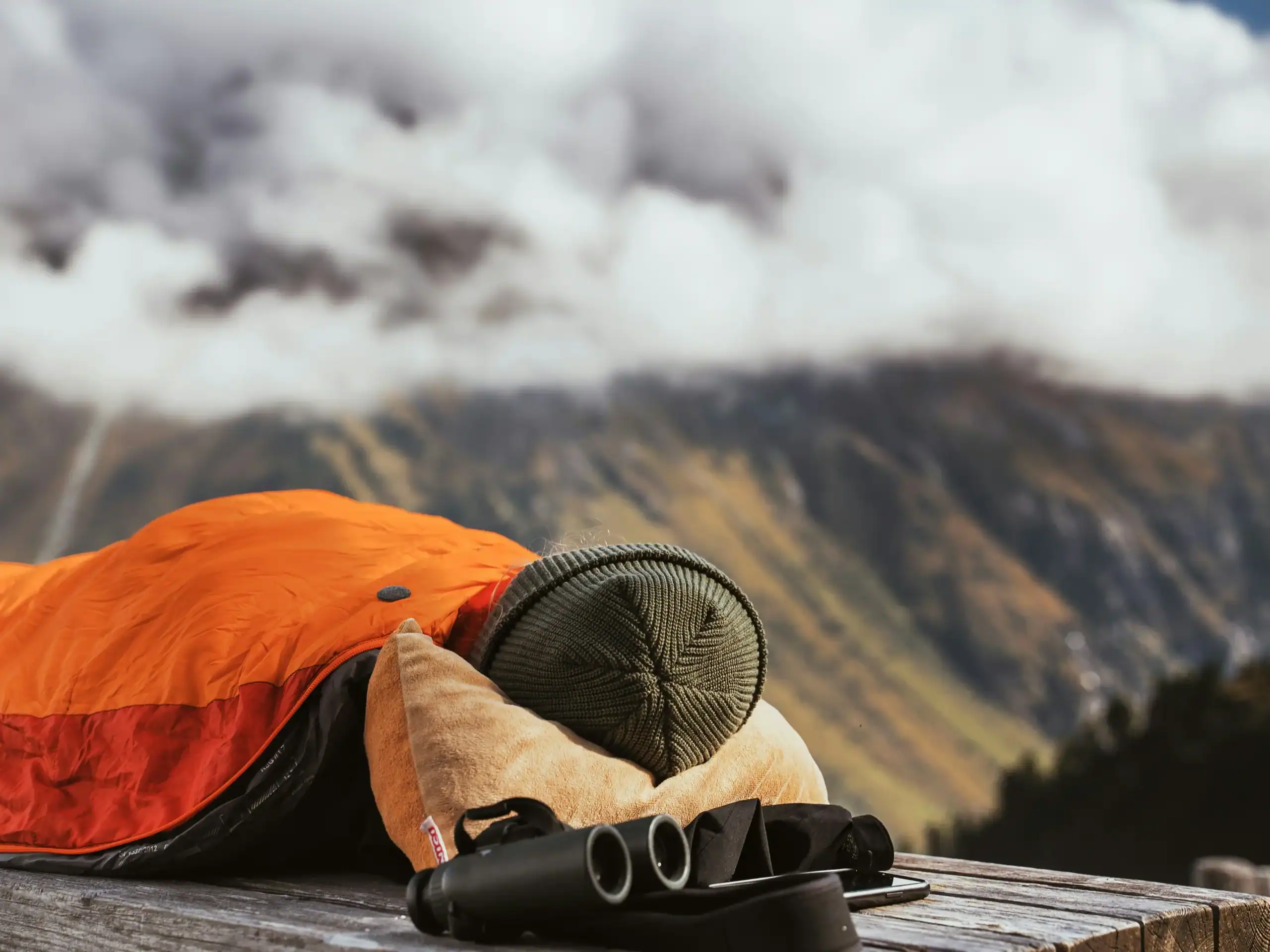 Equipamento de camping ao ar livre com mochila, saco-cama e binóculos sobre superfície de madeira.