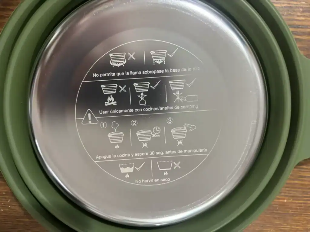 Retail Camp Stove Margin: Charcoal vs Gas 3 Bottom of a camping cookware pot with engraved safety instructions in Spanish, placed on a wooden surface.