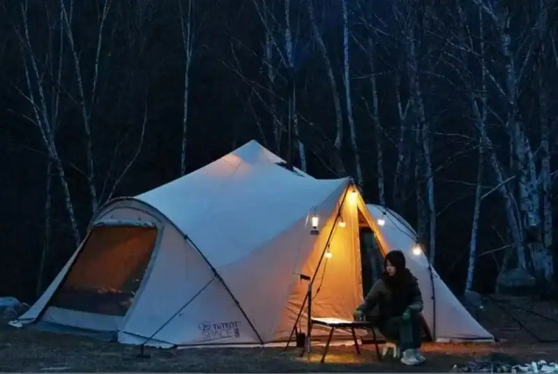 Camping tent illuminated at night in a forest with a person sitting outside.