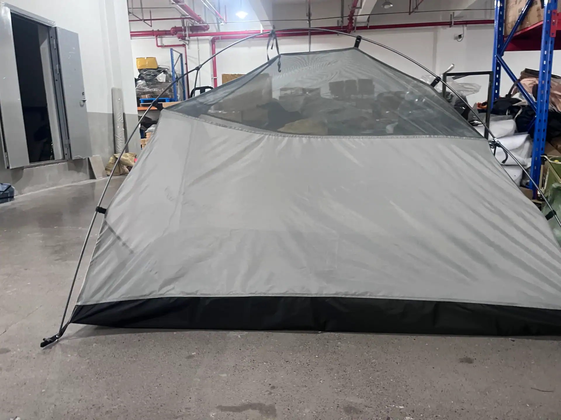 Gray camping tent prototype assembled in an indoor warehouse with shelves and supplies in the background.