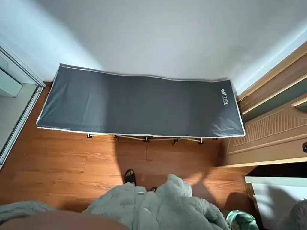Camping Cot Safety Standards: Avoid $50k Compliance Failures 1 A foldable camp bed with a grey cover set up indoors on a wooden floor, with a person standing nearby.