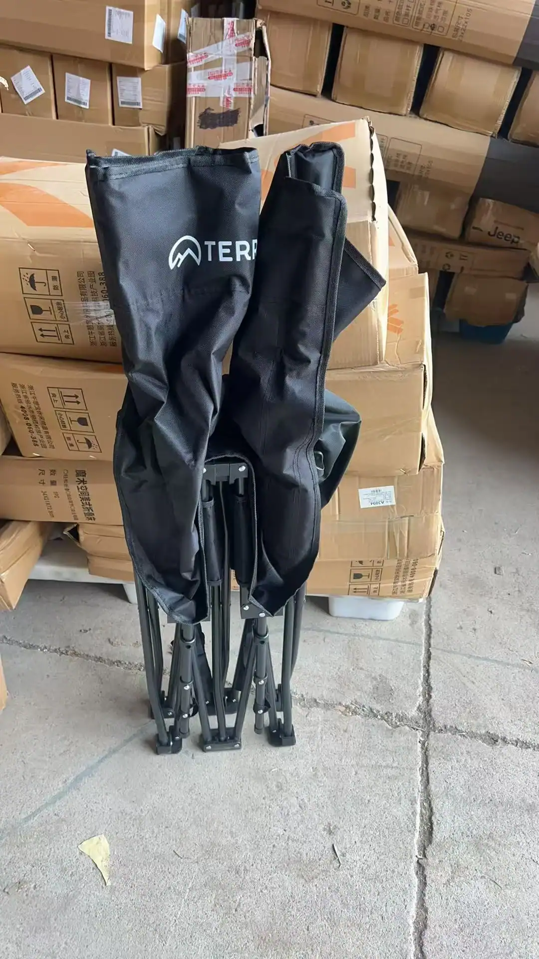 A black folding camp chair in its carrying case positioned in front of stacked cardboard boxes in a warehouse setting.