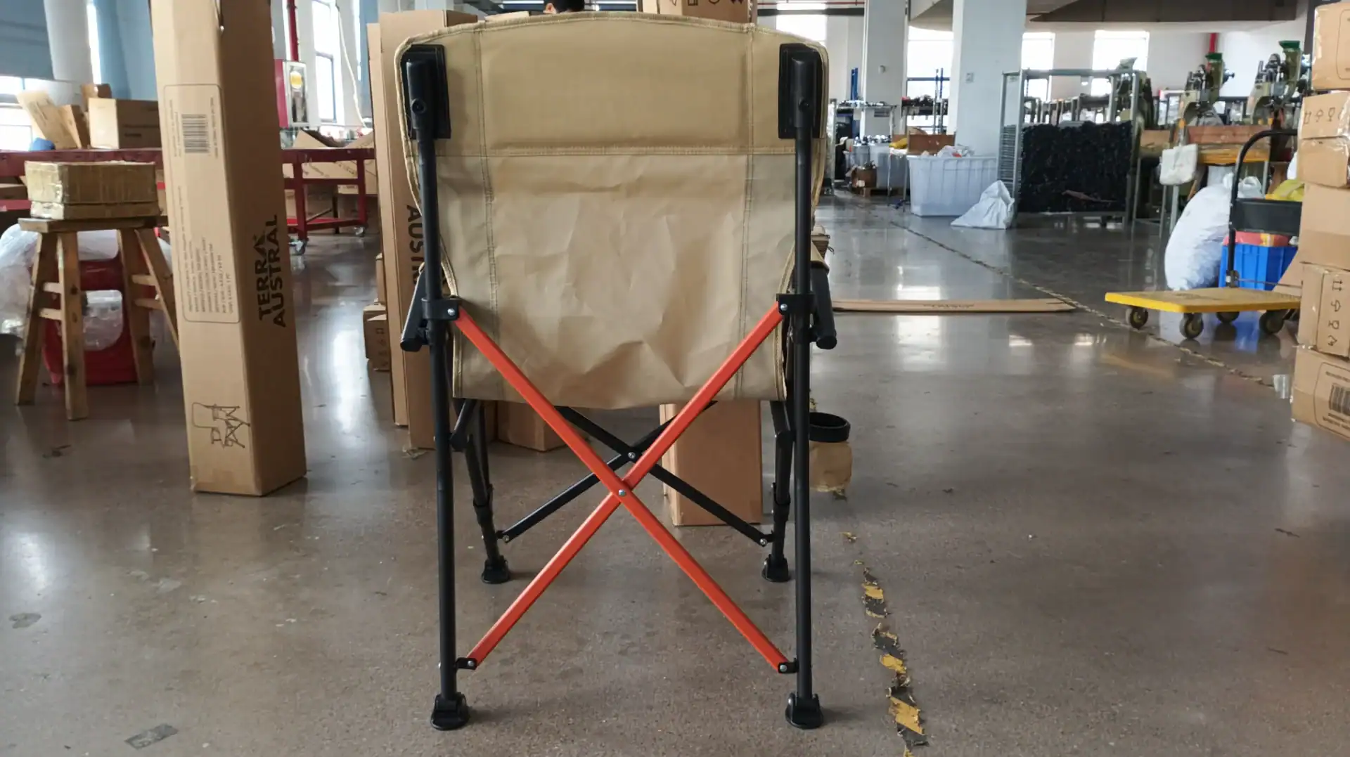 Folded camping chair with tan fabric and orange crossbars in an assembly and packaging facility.