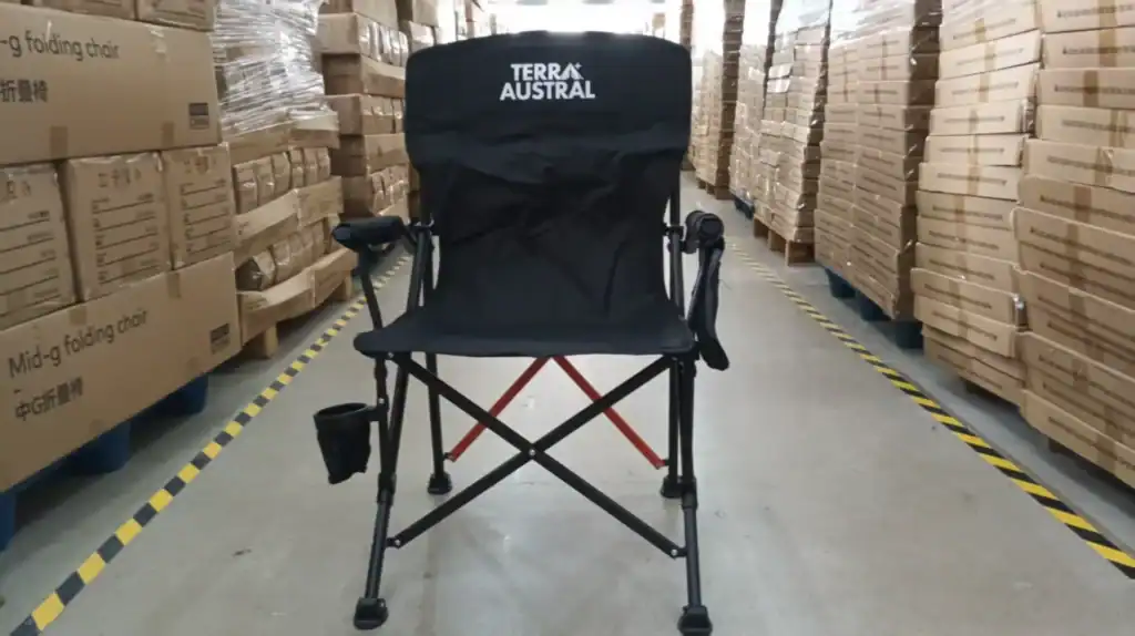 Private Label Camping Chairs: Branding Customization That Lasts 2 Black folding camping chair with cup holder and sturdy frame displayed in a warehouse with stacked cardboard boxes.