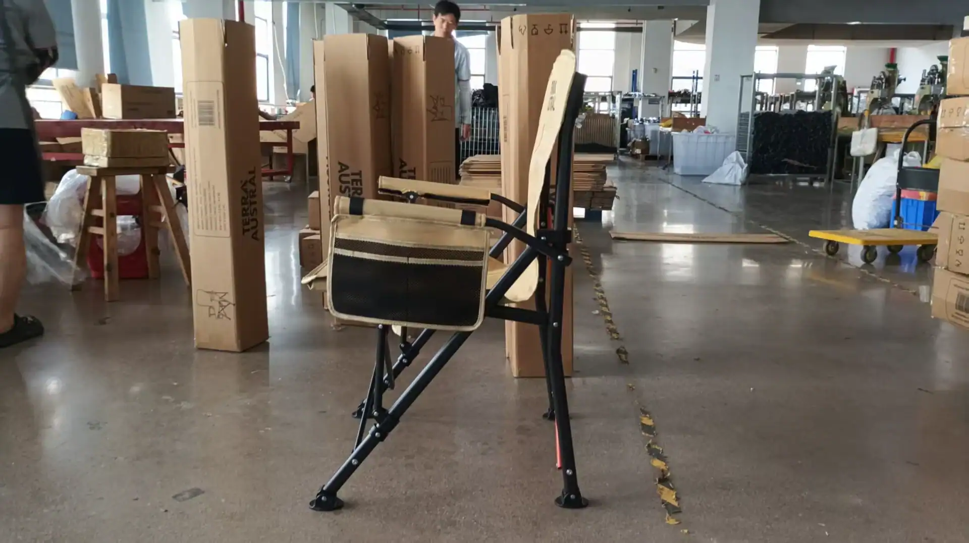 A folding chair with beige seat and black frame in a warehouse surrounded by Terra Austral packaging boxes and assembly materials.