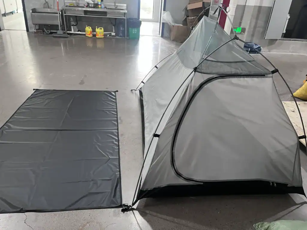 Gray camping tent with a matching floor protector laid out on a polished indoor floor in a workshop or testing environment.