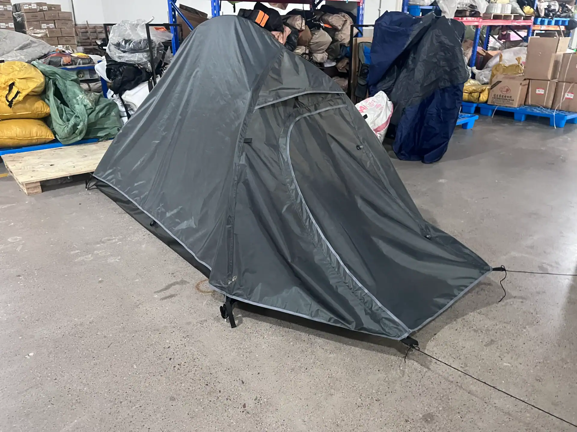 A gray camping tent set up in a warehouse with boxes and supplies in the background.