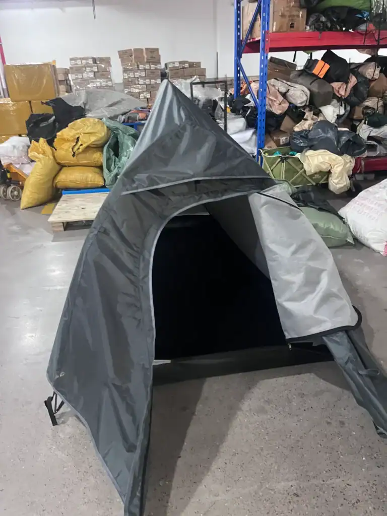 Grey camping tent with an open entrance, displayed in a storage facility with packed items and shelving in the background.
