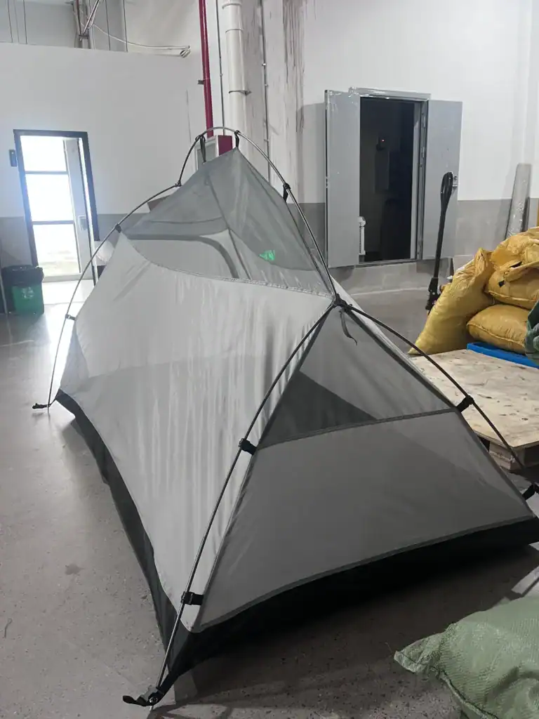 Lightweight gray camping tent with mesh panels set up inside a factory environment with industrial elements visible.