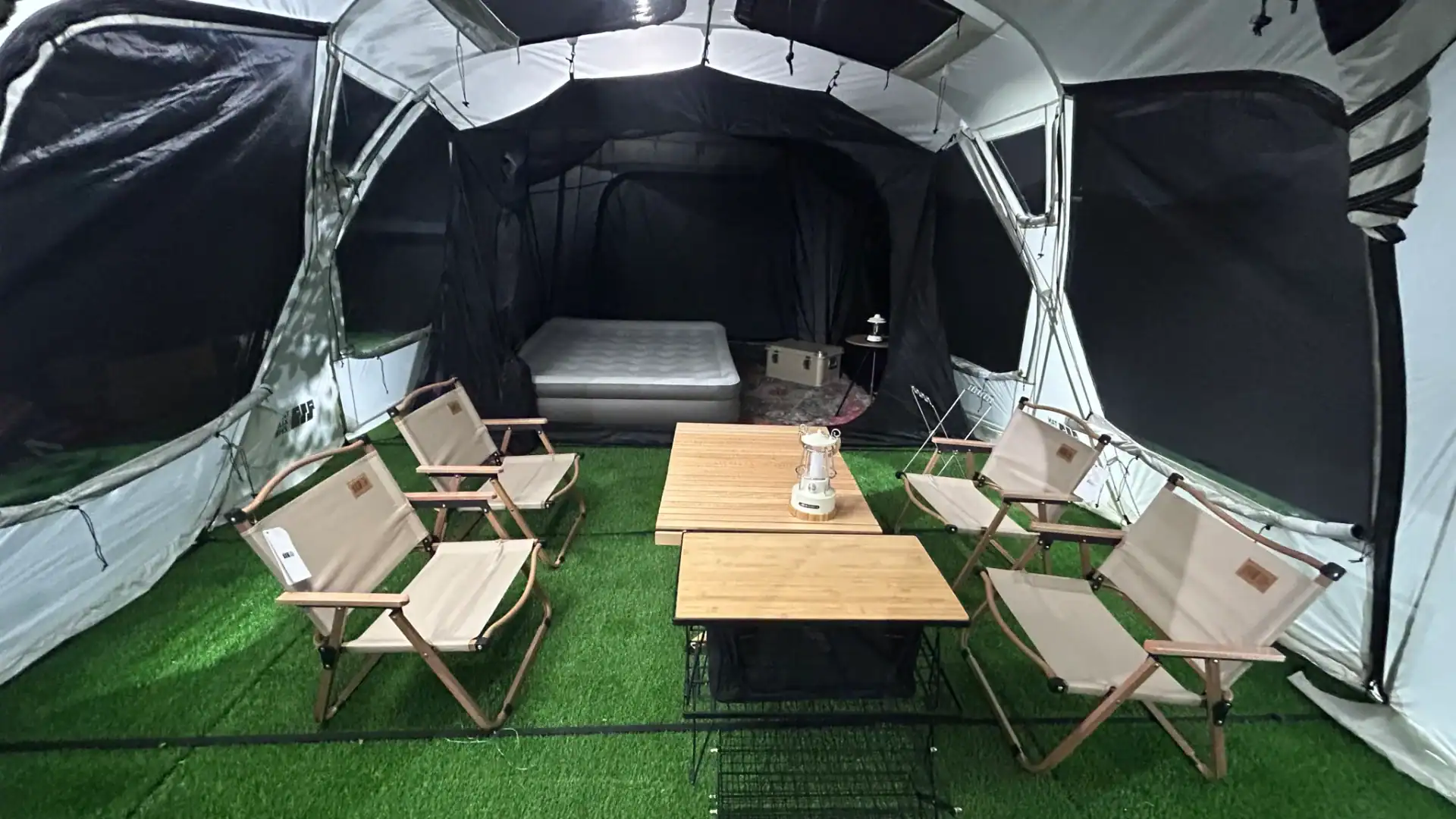 Outdoor camping tent with chairs, table, and mattress on artificial grass.