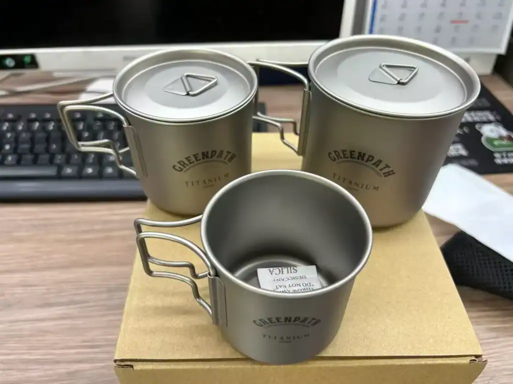 Three Greenpath titanium camping mugs on a cardboard box on a desk.