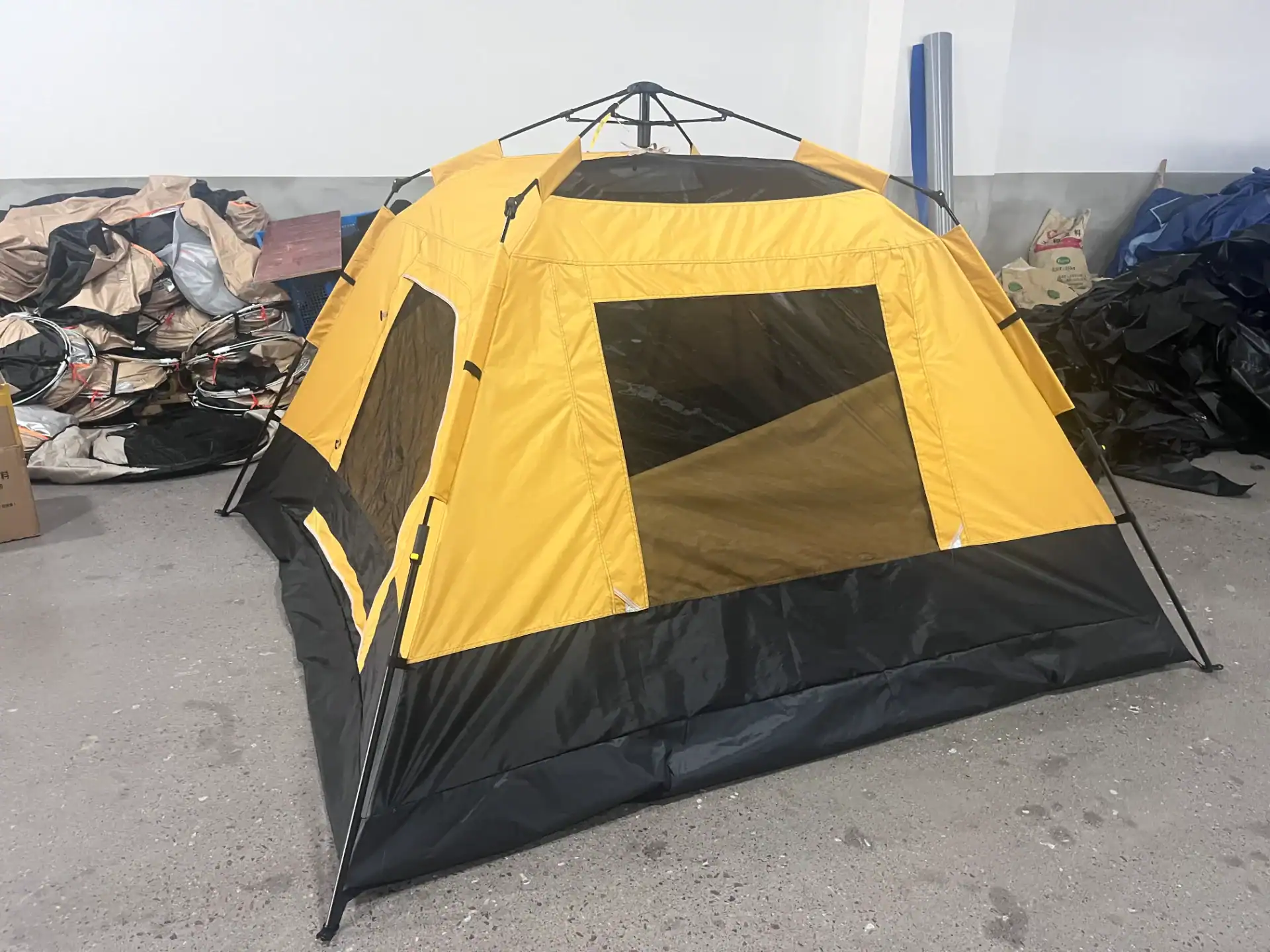 Bright yellow camping tent with mesh windows and black detailing set up in a factory environment with production materials around.