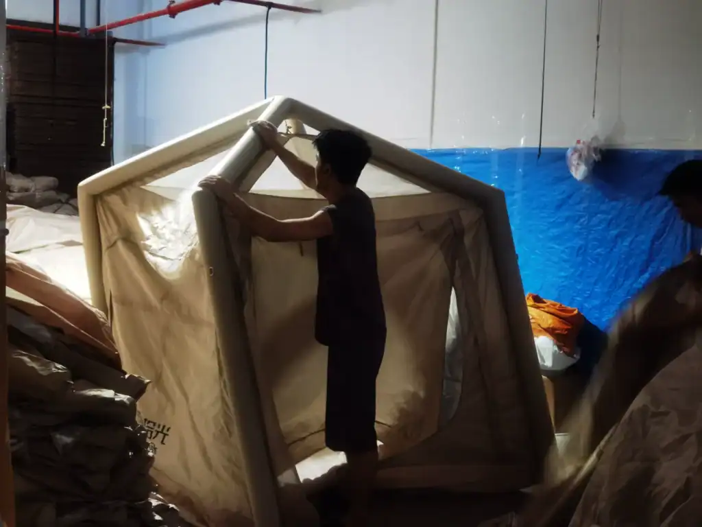 Worker setting up an inflatable camping tent in an indoor workspace with camping materials and tarpaulin in the background.