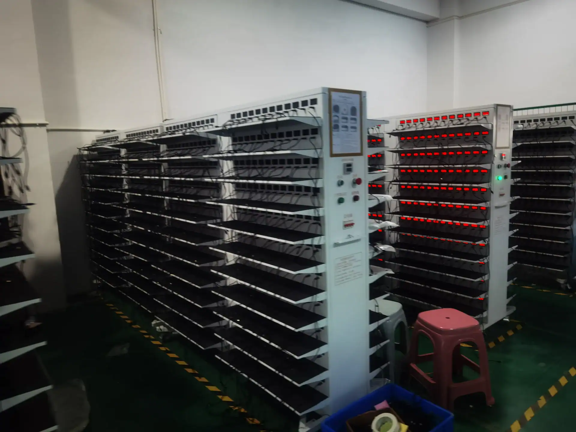 Multiple rows of battery testing racks with connected wires and red LED indicators in a laboratory facility.