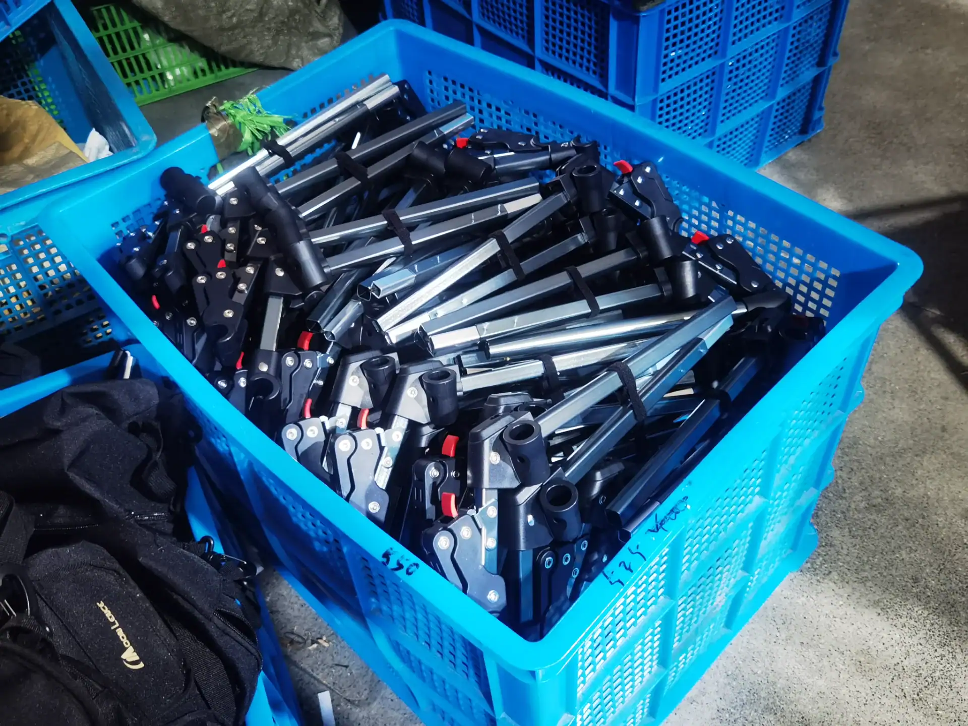 Air Tent vs Pole Tent: Which Survives 35 mph Wind? 17 Blue plastic crate containing numerous metal tent pole assemblies with black and red locking mechanisms, placed on a concrete floor.