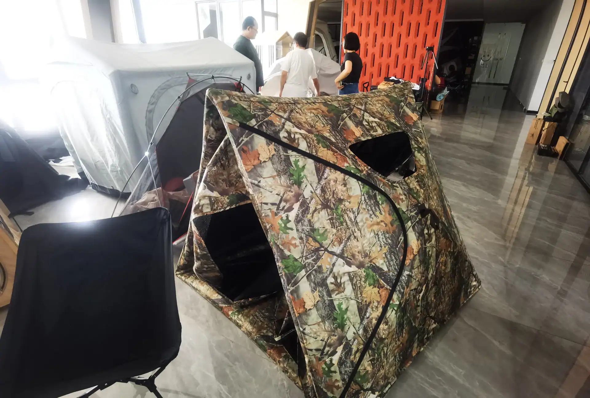 Кемпинговая палатка с камуфляжным рисунком, выставленная в помещении рядом с другим туристическим снаряжением, на заднем плане три человека осматривают другую палатку.