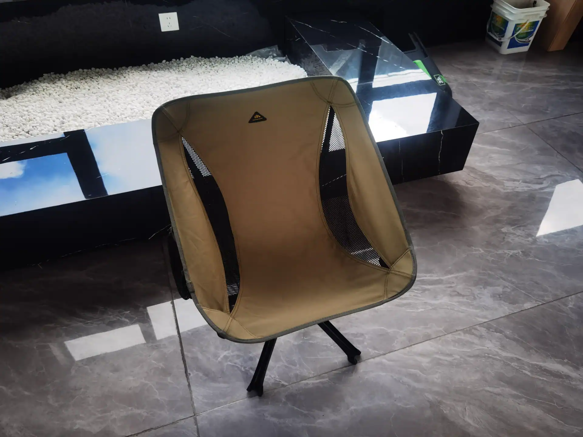 Folding Wagon Packaging Drop Test: The 60cm Export QC Rule 148 A tan-colored lightweight outdoor folding chair with mesh sides, placed on a tiled floor near a black marble structure.