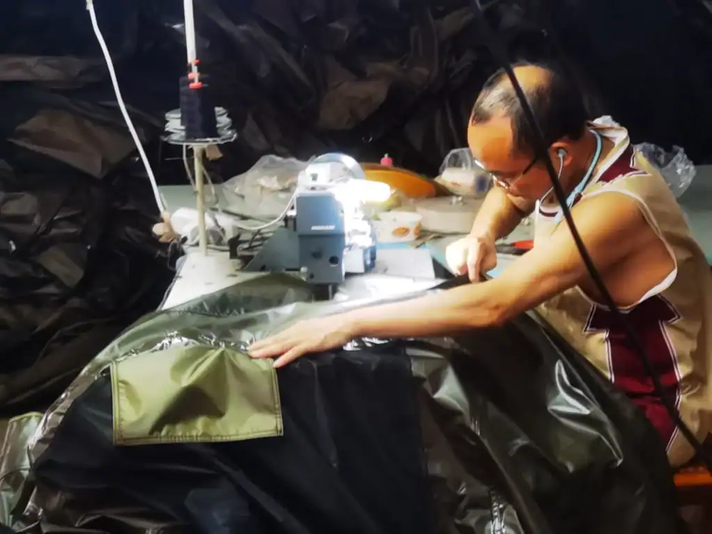 A craftsman working on stitching durable camping tent fabric at a sewing station with a sewing machine, surrounded by black and green materials, in a workshop setting.