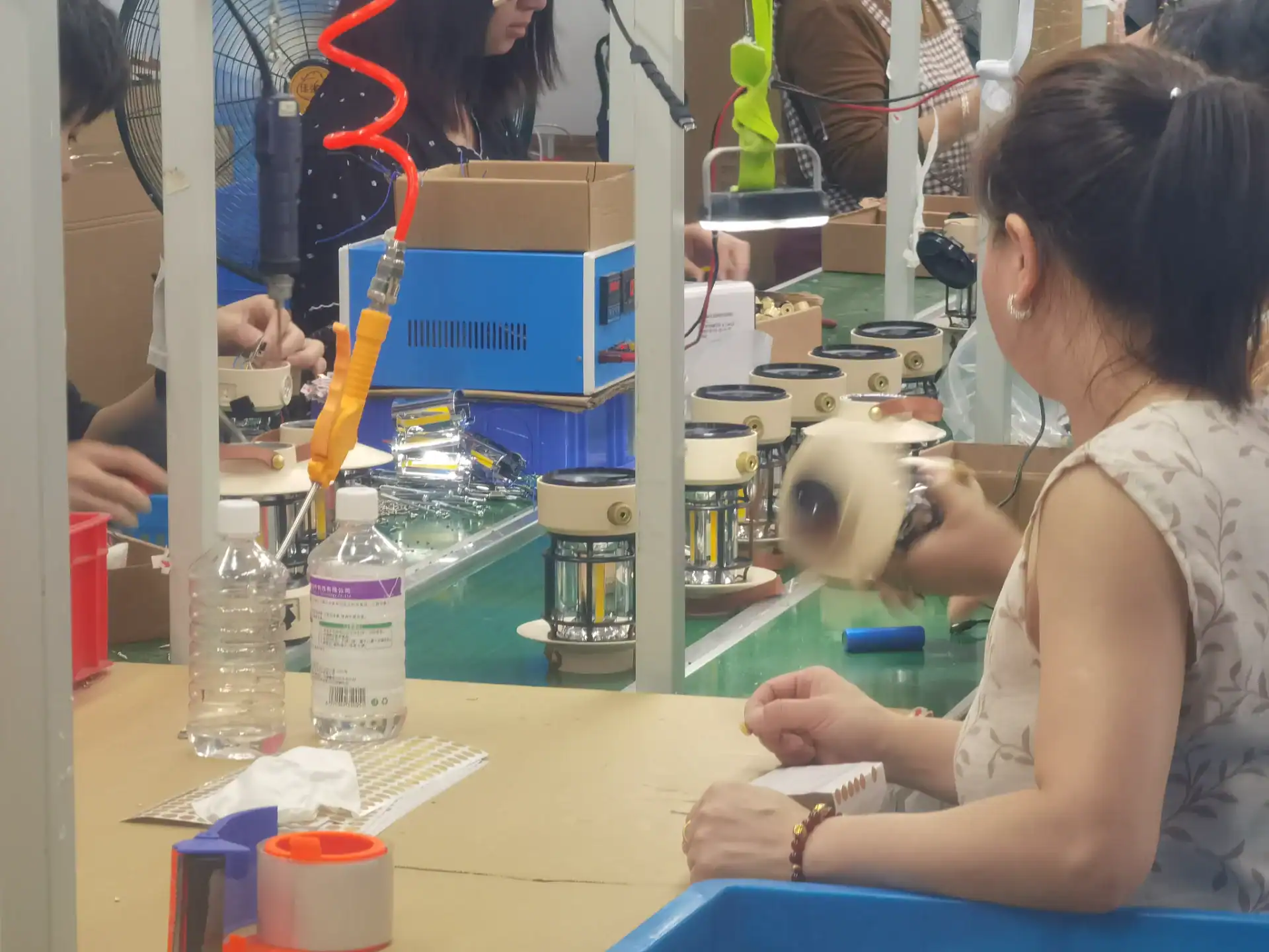 Workers assembling components on a production line in a factory, with tools, assembled devices, and materials on the table.