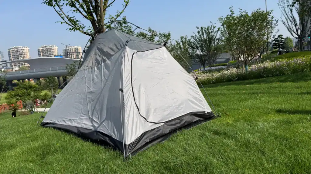 A gray camping tent set up outdoors on a lush green lawn in a park with trees, flowers, and modern buildings in the background.