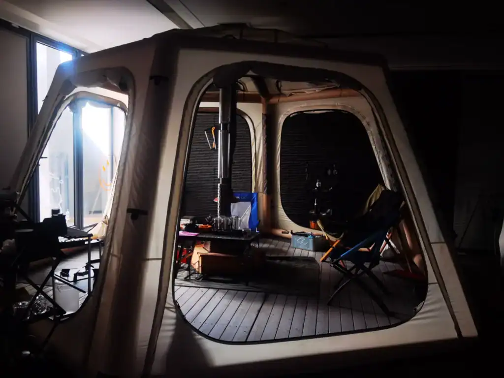 Ein geräumiges beiges Campingzelt, das im Innenbereich aufgebaut ist und Stühle, einen Holzofen, Beleuchtung und weitere Campingausrüstung auf einem Holzboden zeigt.