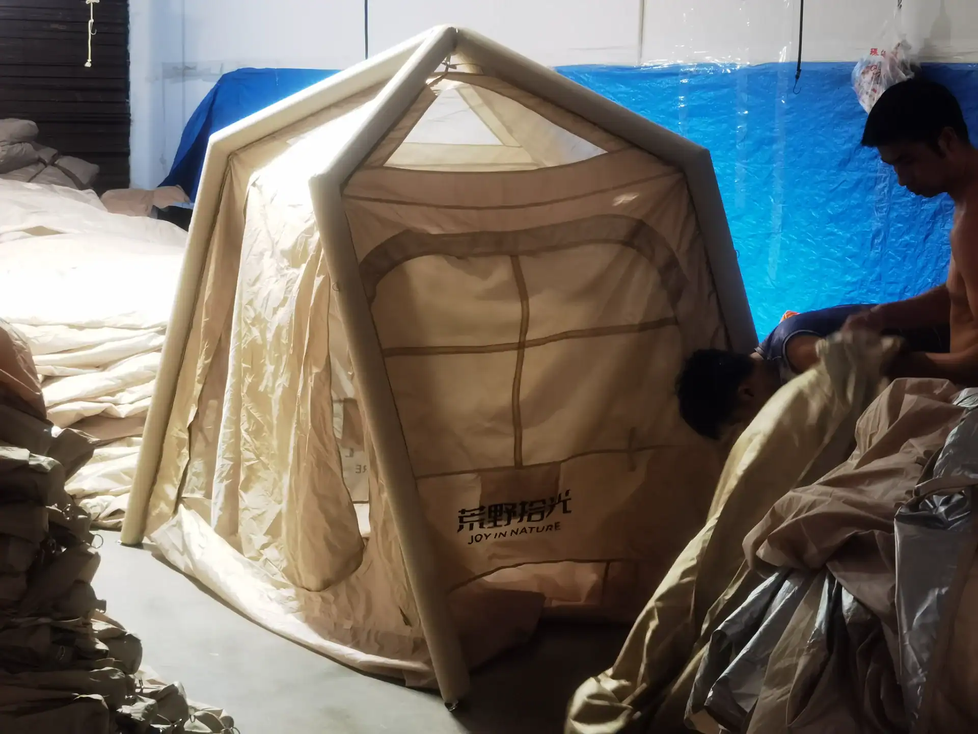 Workers assembling an inflatable camping tent in a workshop, surrounded by fabric materials and tools.