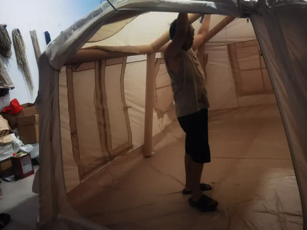 Custom Inflatable Tent Case Study: 45-Day Retail Launch 1 A man assembling a large beige camping tent indoors with visible structural poles. Various materials and boxes are in the background.