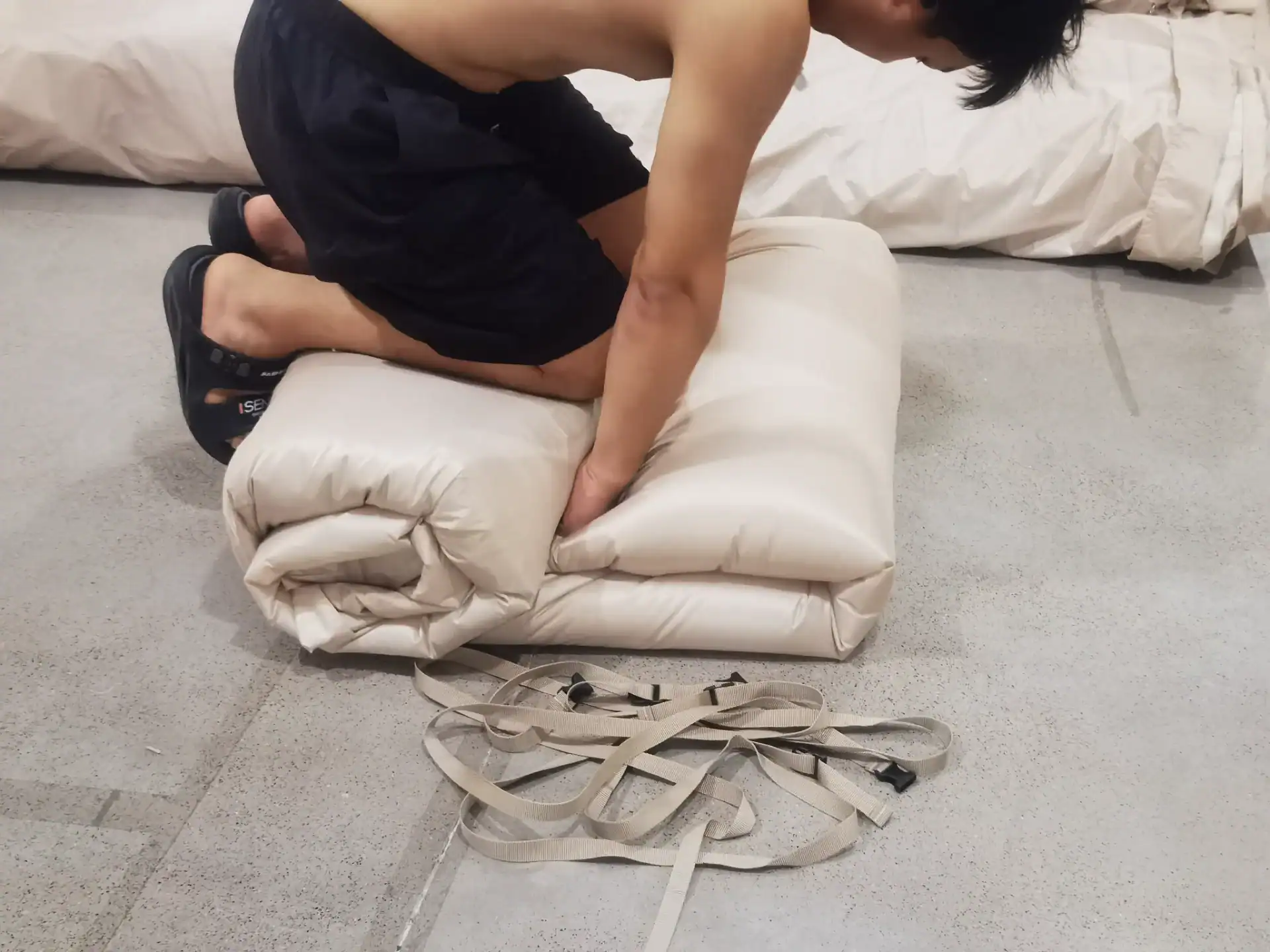 ベージュのキャンプ用寝マットの上にひざまずいて丸めている男性。ストラップがコンクリートの床に置かれている。.