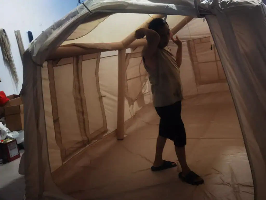 Ein Mann testet ein geräumiges beige Campingzelt in einem Innenraum; er steht darin mit erhobenen Armen, umgeben von Aufbewahrungsgegenständen.