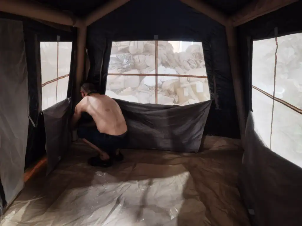 A person setting up the interior of a large camping tent with visible windows and outdoor materials in the background.