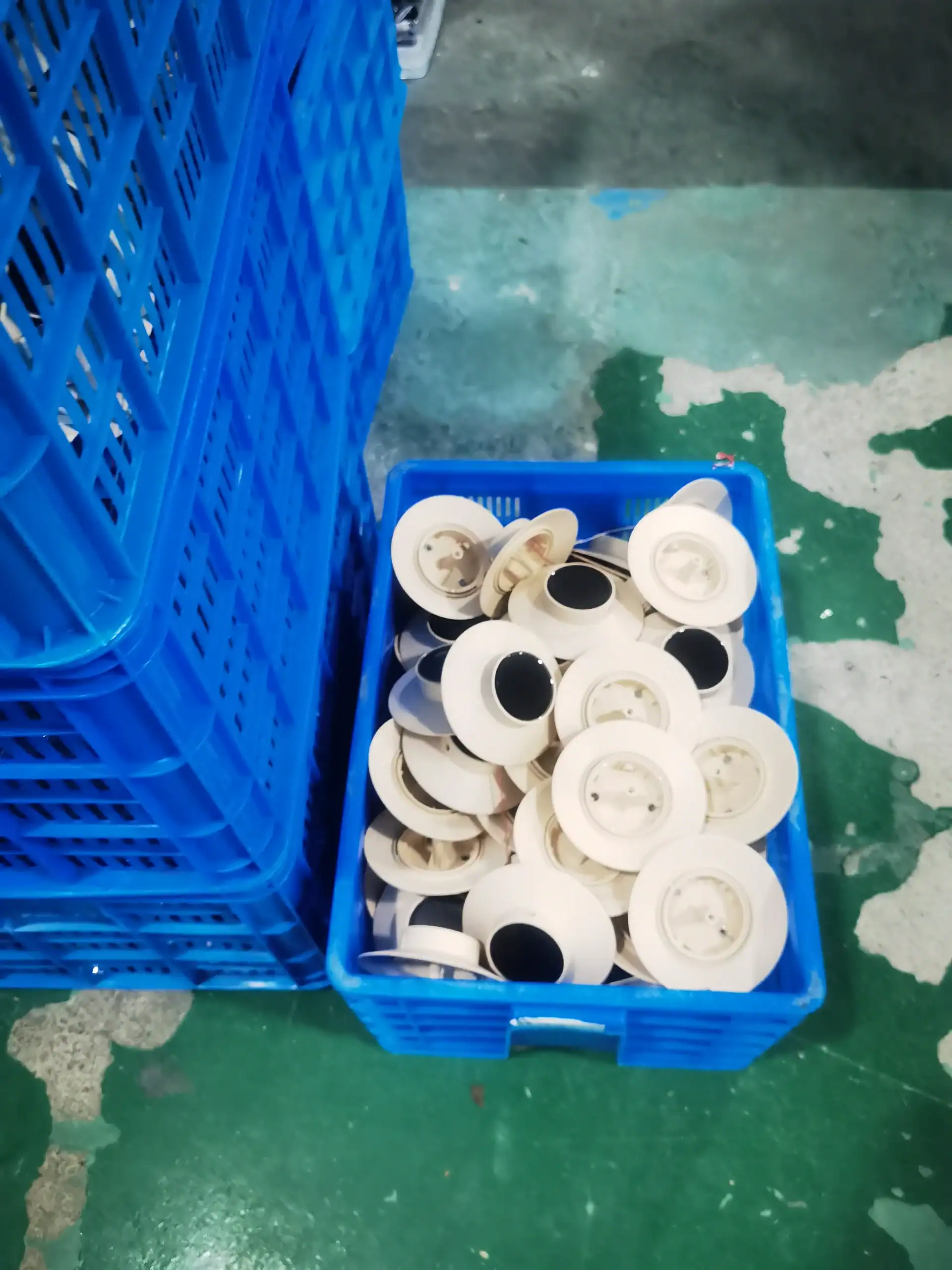 Close-up of white plastic circular parts stored inside a blue crate in an industrial facility with a green concrete floor.