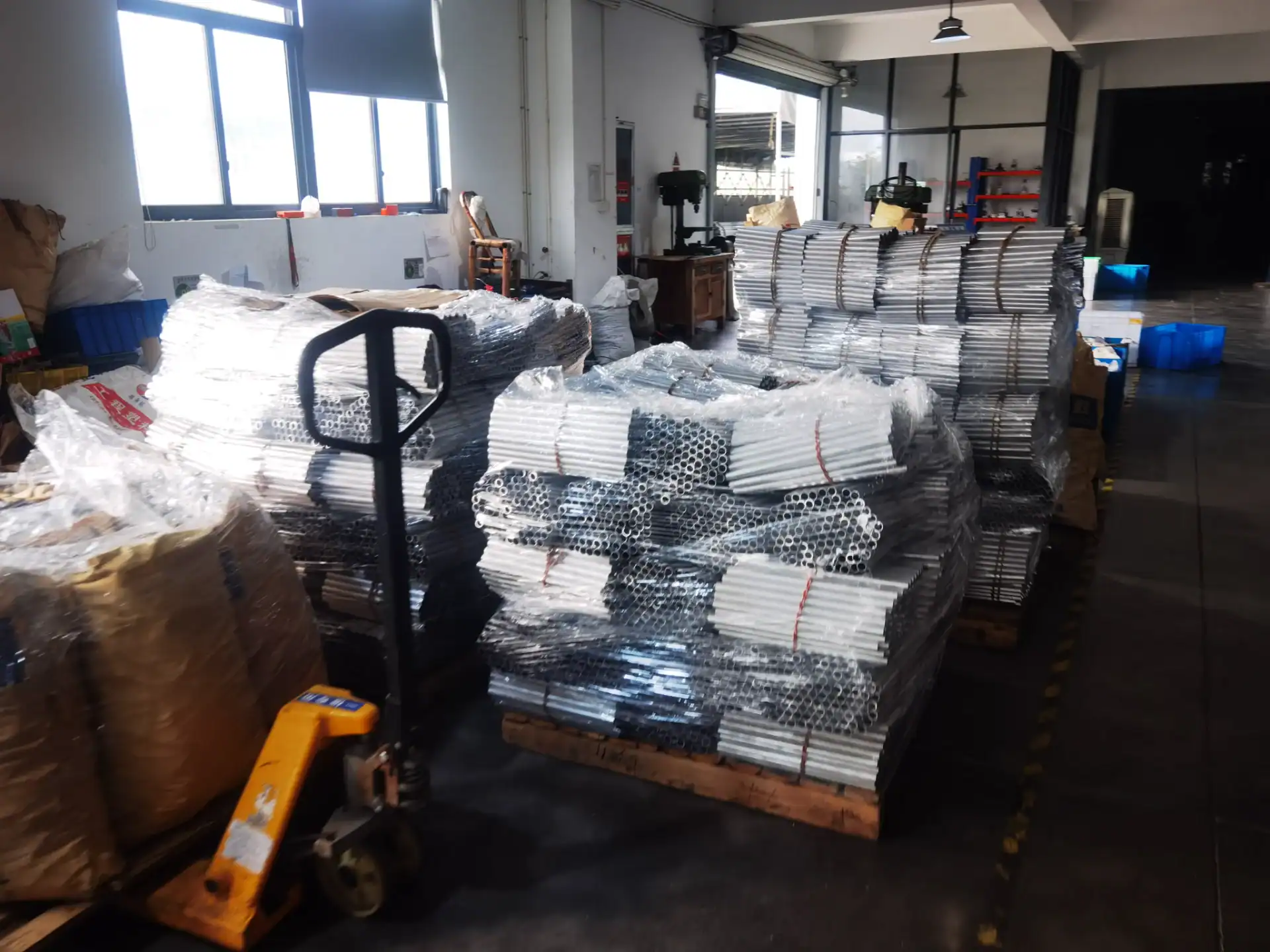 Stacked metal tubes wrapped in plastic and bound with straps on wooden pallets in a warehouse with light streaming through windows.