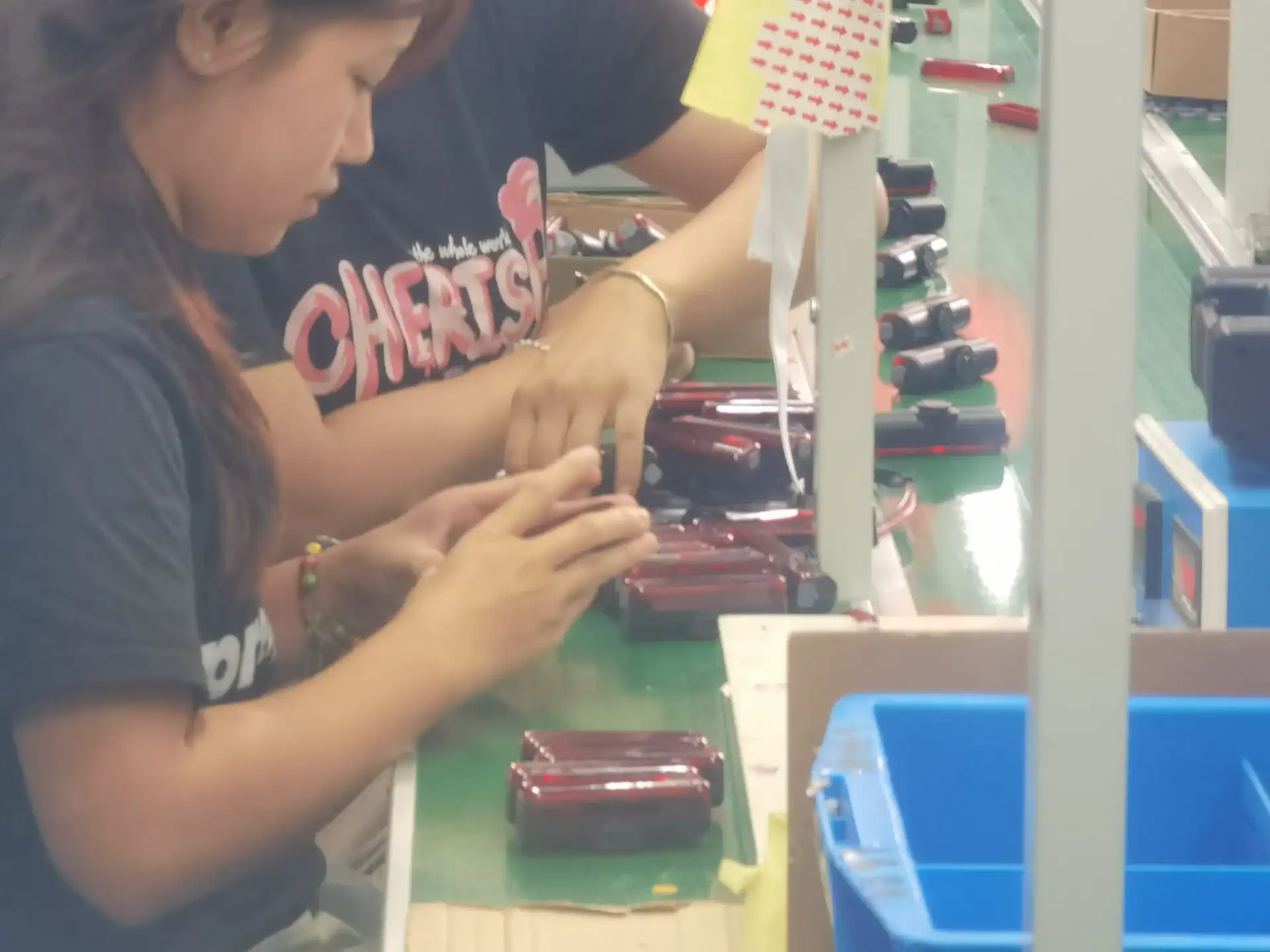 Inflatable Tent Manufacturer Audit: Seams, Valves & Tubes 129 Two workers assembling small handheld devices at a workstation in a factory with tools and equipment on a green surface.